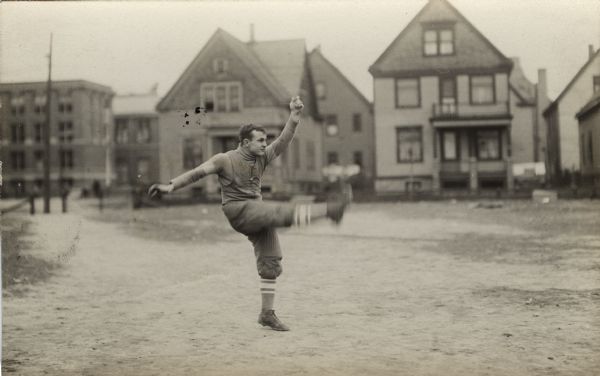 Side view of a man presumably kicking a football. He is in a lot, and houses are in the background.