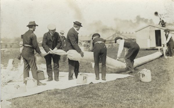 Men working outdoors to fill what appears to be sand bags. Some of the bags are marked with a "P." There are industrial buildings in the background.