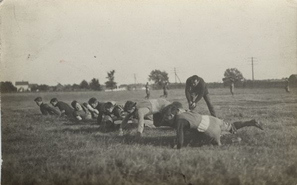 Side view of men in a line in a field. They appear to be doing push-ups, and one man stands behind them perhaps coaching or timing them.