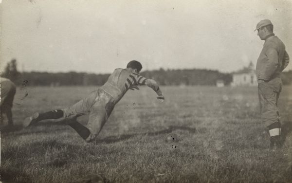 A football player in a field is leaping in mid-air towards a football in the grass. A man stands watching nearby, perhaps a referee or coach.