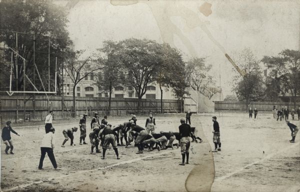 Elevated view of men playing football on a field. There is a large wood fence in the background, and buildings beyond.