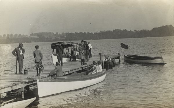 View from shoreline of a group of people standing on a pier. Three people are in a boat along the side of the pier, and another boat with a roof is on the other side. In the far distance is the opposite shoreline.