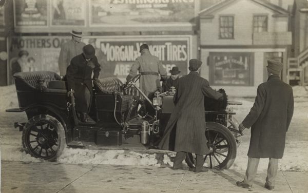 View from sidewalk of men dressed in hats and jackets exiting an automobile at the curb. There is snow along the curb, and across the street are billboards.
