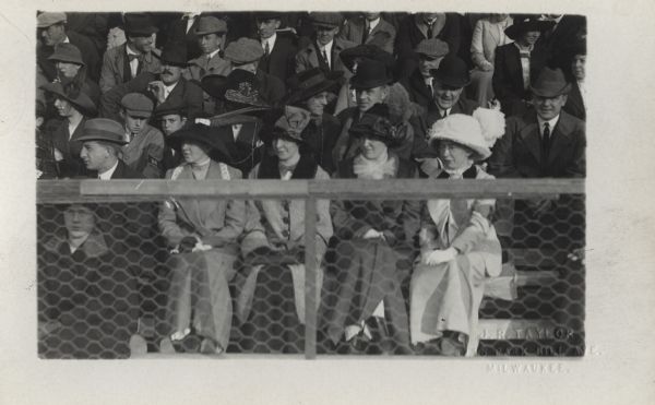 View over fence of four women wearing hats and coats sitting among a crowd of spectators.