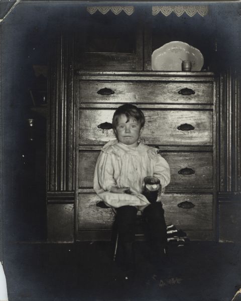 Unidentified young boy sitting on a chair in front of a dresser.