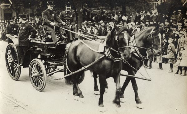 Four officers are riding in a carriage labeled "Fire Department" pulled by two horses. They appear to be part of a parade. There is a crowd of people standing along the street in the background.