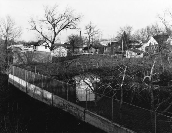 Elevated view of backyards in a neighborhood looking over trees, a shed, and a fences. In the background are houses and a smokestack.