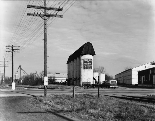 View over roads and railroad tracks of industrial silos for the Brunsell Co. Trucks are parked nearby, and warehouses are on the right.