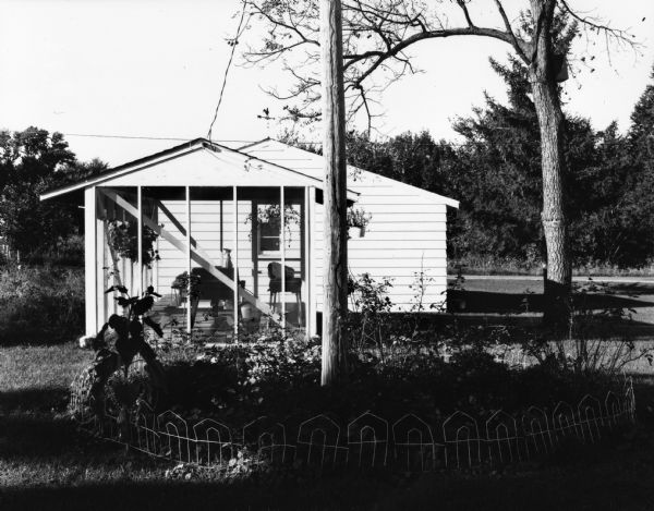 Small house with screened-in porch. In the foreground is a fenced-in garden surrounding a telephone pole.