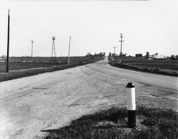 A short black and white pole marks where a road converges with a state highway that stretches to the horizon. In the mid-distance before a hill is a railroad crossing. There is a windmill on the left, and a farm in the background.