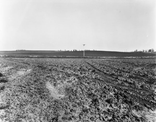 View over plowed field, with a telephone pole in the middle distance. Farm buildings are on the hill in the background.