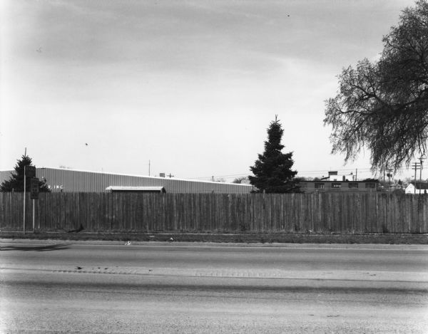 View across stateline road towards a wooden fence lining the road on the opposite side. A few trees and buildings can be seen over the fence.