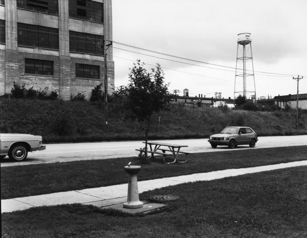 View across road, perhaps Pleasant Street, of industrial buildings. In the foreground is a drinking fountain and picnic table. Two cars are parked nearby.