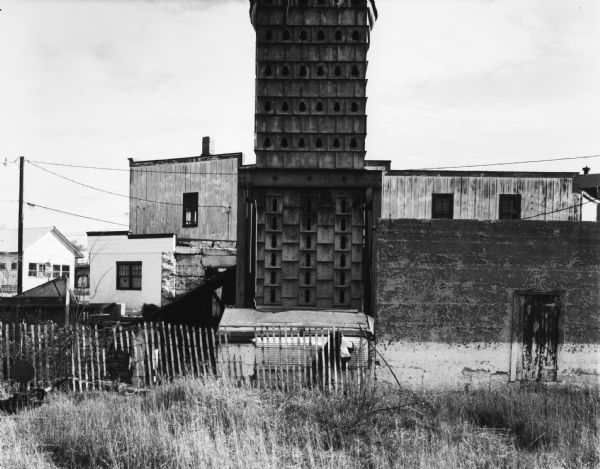 View of a tall wood structure with rows of holes, perhaps a pigeon house. A cyclone fence is in front of it, and behind are industrial buildings. In the far background on the left is a sign for a grocery store above an ice machine.