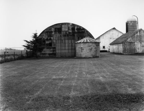 View across lawn of a Quonset hut-style building and metal grain bin. A barn and other farm buildings including a silo are on the right. To the left is a fence bordering a field with an irrigation system on it.