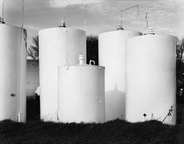 Five large cylindrical tanks. Trees and an industrial metal building are in the background.