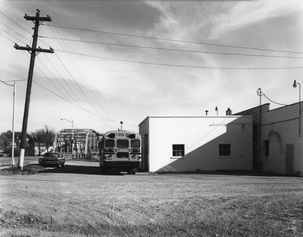 View across parking lot of a school bus and a car parked near a building on the right. In the background there is a metal bridge with trees and houses behind it.