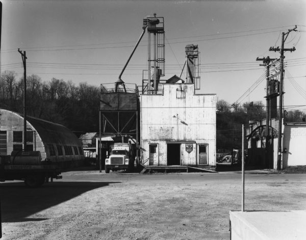 View of large building which is depositing feed into a truck docked on its left. On the left there is a Quonset hut-style corrugated metal warehouse, and on the right is a brick industrial building.