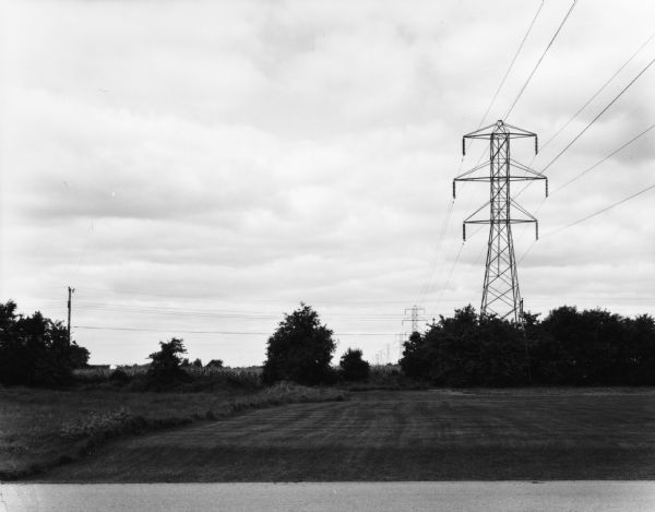 View across small section of road (Huebbe Parkway?) towards a mowed field. A large power or telephone tower rises above a group of trees, and more towers are in a line behind it stretching towards the horizon.