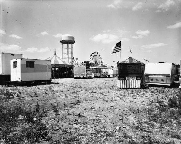 View of carnival with a water tower in the background. There are trailers, and a carousel is on the left. Carts selling food like popcorn and sausage are parked nearby. An American flag flies from a cart marked "Redwood Sign Shop." A Ferris wheel and other amusement rides are in the far background.