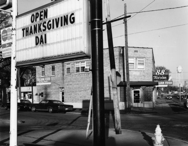 A sign prominently displays "OPEN THANKSGIVING DAI" in the top left corner of the frame. Across Third Street behind the sign is the 88 Tavern. Down Portland Avenue in the background a newer metal water tower and just to its right is the old stone water tower.