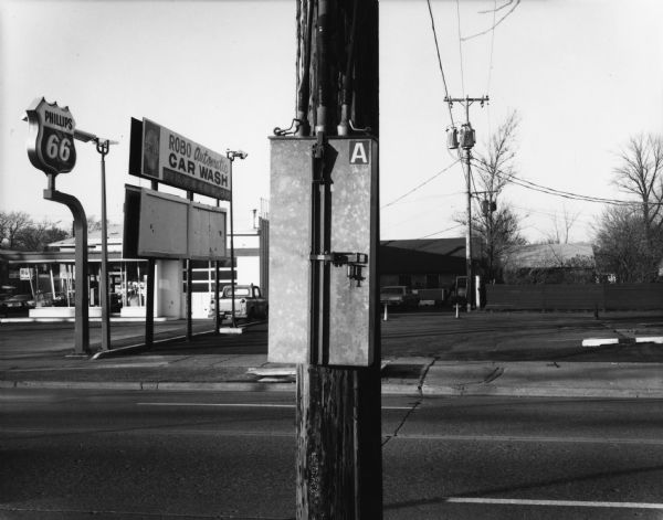 An electrical box on a wooden pole is in the center foreground. Behind it is a street, and on the other side of the street is a Phillips 66 service station and a ROBO Automatic CAR WASH.