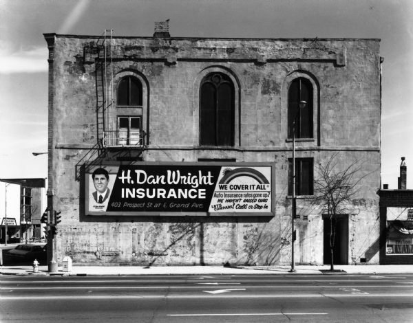 View from across street of a rectangular dilapidated building featuring arched windows. A billboard for H. Dan Wright Insurance is under the fire escape.