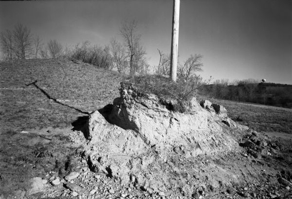 A telephone pole emerges from a dirt mound and casts a shadow across the land. Trees and a water tower can be seen in the distance. Bible Camp Road.