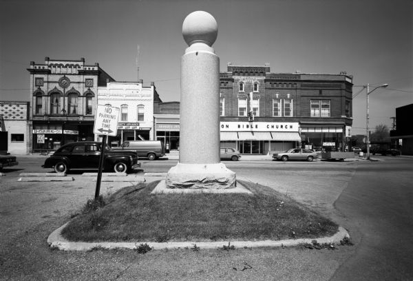 Monument on grassy area. Behind are storefronts.