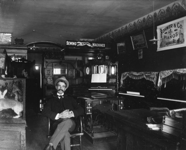 A well-dressed man sits in a chair in a Schwaller Music Store on Pine Street. Various pianos are on display. There are signs for Sohmer Pianos and New Home Sewing Machines. There is also a glass case of taxidermied animals.
