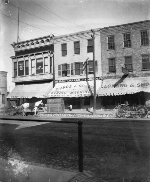 Looking east across Pine Street at the F. A. Schwaller Music Store / Schwaller's Real Estate Office, flanked by Theodore Riel's dry goods and grocery store in the Florence block (left) and Jacob Wien¿s Chicago Store, which sold clothing and shoes, in the Haas building (right). A horse and carriage are in front of the Riel store and a piano crate is in front of the music store. A man and young child are in the doorway of the music store.  In 1904 Riel sold his stock to Jacob Wien, who moved his business into the former Riel space in August 1904.
