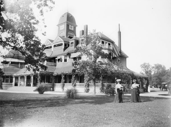 Two women pose on the lawn in front of a large estate. The building, with a large, wrap-around porch, and clock tower, was probably used as a resort or hotel. In the background on the drive is a horse-drawn carriage.