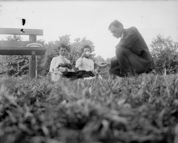 Low angle view from the grass of two women and a man having a picnic near a wooden bench.