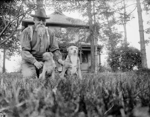 Low angle view of a bearded man kneeling with two dogs in a yard. In the background is a two-story house.