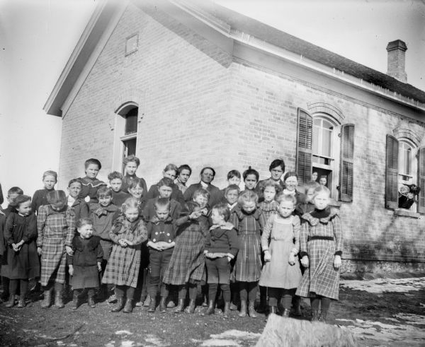 A group of children pose in front of a brick schoolhouse. Several children peer out through the schoolhouse windows on the right.