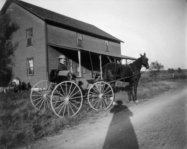 Side view of a man sitting in a carriage pulled by a horse in front of a house. The photographer's shadow falls into the photograph, just in front of the carriage.