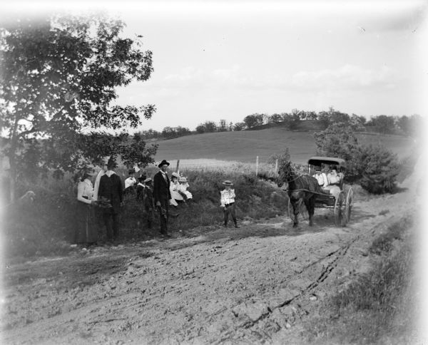 A group of men, women, and children sit in the shade on the side of a country road. A dog is sitting under a tree on the left, and a horse-drawn vehicle approaches the group from the right.