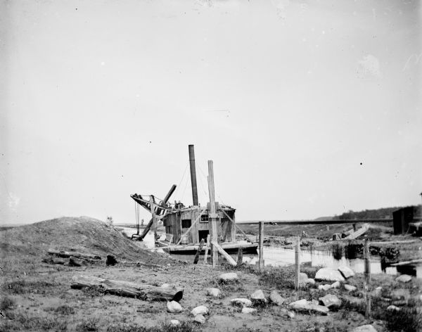 View from shoreline of a dredging barge on a canal. Several large rocks are scattered in the foreground.