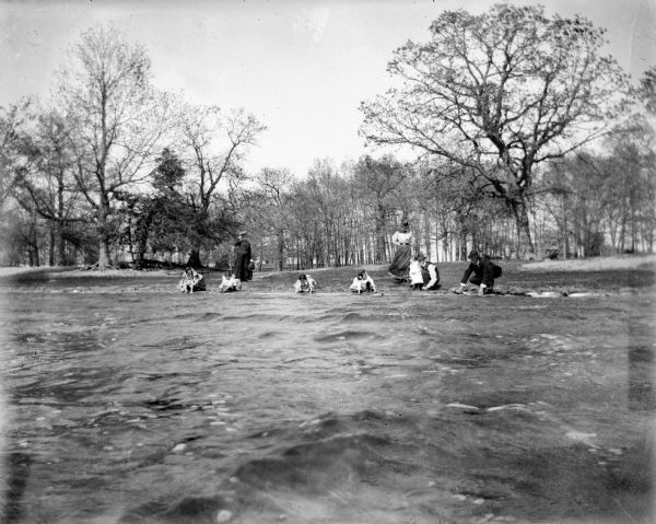 View from water of two men and five young children playing at the edge of a lake. Two women stand nearby and in the background are trees.