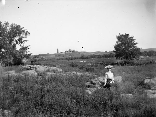 A woman sits in a rocky field wearing a broad-rimmed hat. In the distance, a large building with a tower is visible on the horizon.