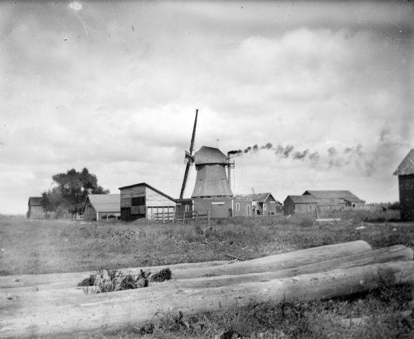 View from field of a cluster of farm outbuildings, including a windmill. Smoke is coming out of a smokestack attached to the back of the windmill.