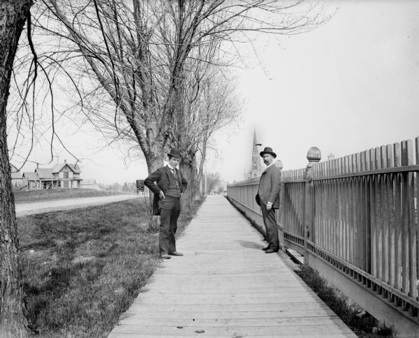 Two men stand on a boardwalk next to a decorative wooden fence. A house and church are visible in the background, and the back of a carriage traveling down the road is behind the man on the left.