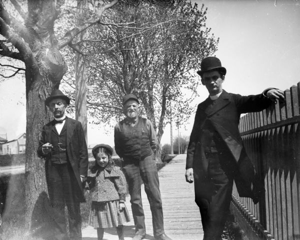 Three men stand on a board sidewalk with a young girl. A decorative wooden fence lines the boardwalk and a house is in the far background.