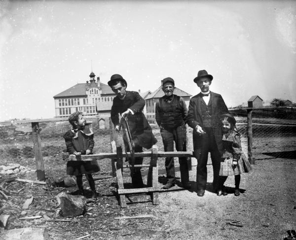 Three men stand with two young girls near a sawhorse. One of the men holds a saw. A large building is visible in the background.