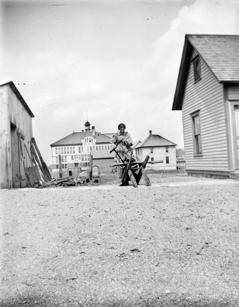 A woman stands at a sawhorse between two small outbuildings using a hand saw with a sawhorse. A large building is visible in the background.