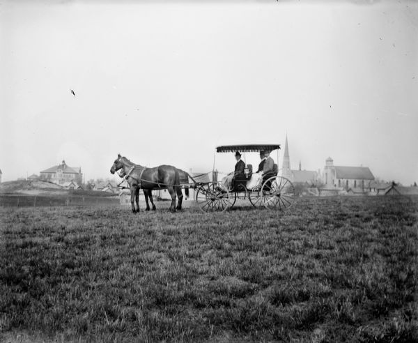 Two men and a woman ride in a horse-drawn carriage across a field. They have blankets over their laps. A town is visible in the far background.