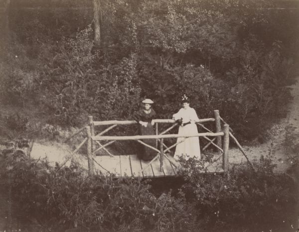 Elevated view of two women, Mrs. Christine Rawsteck(?) and Mrs. J.W. Hommel(?), standing on a rustic bridge. The bridge is a part of a larger path that runs through a wooded area.