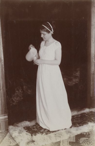 Mrs. G.E. Vandercook stands in a Grecian-inspired costume and pours a beverage from a ceramic pitcher.