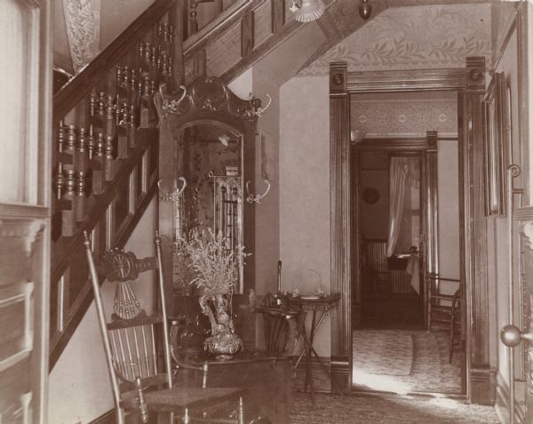 View through open front door of entry of home decorated in the Victorian style, including a carved wood stairway and a hall tree bench with a beveled mirror. Elaborate rugs and decorative wallpaper adorn the house, in addition to the carved woodwork and wooden furniture. The home was owned by a Mrs. Daffo and located on North Henry Street.