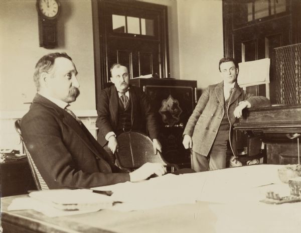 Three men, identified from left to right as Mr. Schildhauer, Mr. Parker, and Mr. Potter, at the Internal Revenue Office. The men are gathered around a large table.